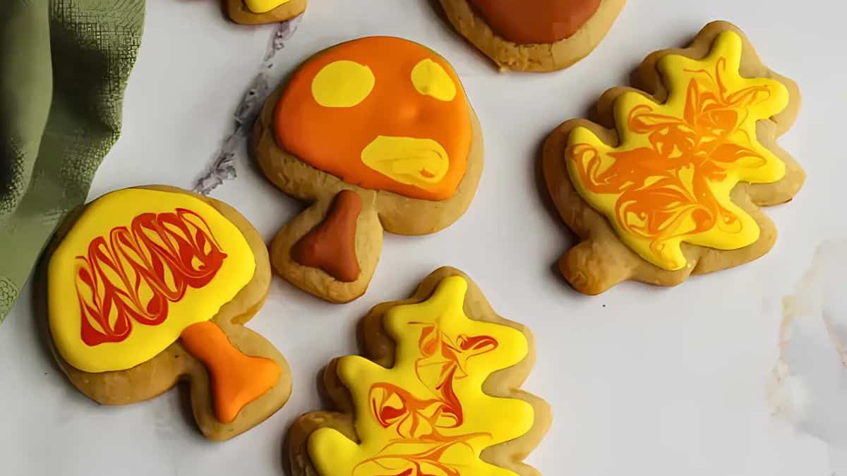 Cookies shaped like mushrooms and autumn leaves, decorated with orange, yellow, and brown icing. They are arranged on a marble surface with a green cloth nearby.