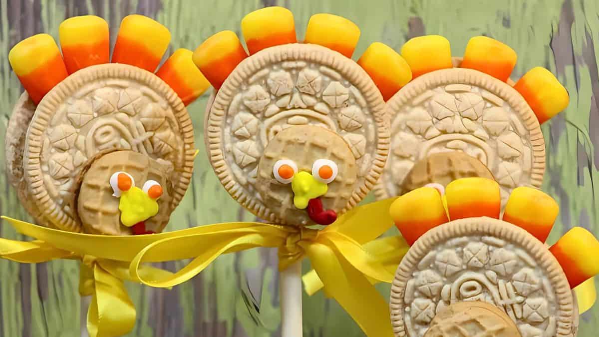 Festive turkey-shaped cookie pops with candy corn feathers, icing eyes, and beaks. They are tied with yellow ribbons, displayed in front of a painted green backdrop.