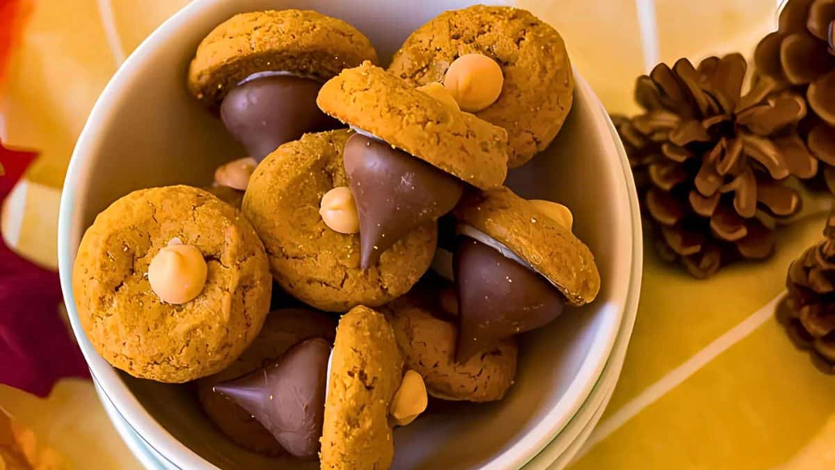 A bowl filled with acorn-shaped treats made from cookies, chocolate kisses, and butterscotch chips. The bowl is surrounded by a decorative autumn setting with pinecones and leaves.