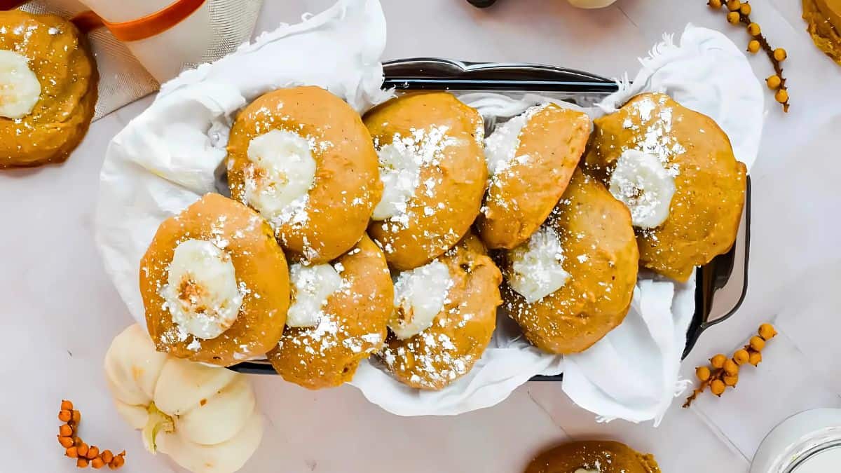 A basket filled with pumpkin cookies topped with icing and powdered sugar, set on a white cloth. Mini pumpkins and orange berries are scattered around for decoration.