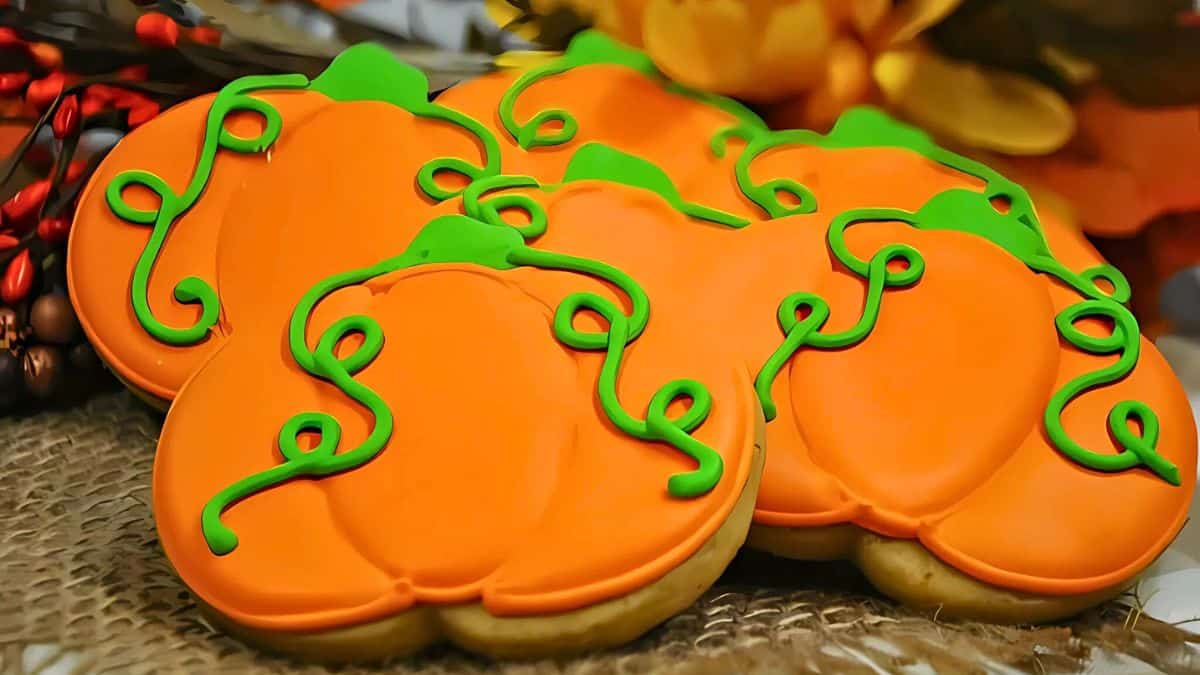 A group of cookies decorated to look like orange pumpkins with green stems and vines, arranged on a burlap surface. Bright flowers and berries are in the background.