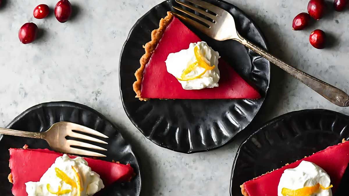Three slices of vibrant red cranberry tart on black plates, each topped with whipped cream and lemon zest. Silver forks are placed on the plates, and fresh cranberries are scattered on the gray marble surface.