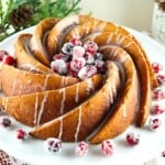 A spiral-shaped Bundt cake with icing drizzles, garnished with sugared cranberries, displayed on a white plate. Pine branches and pinecones decorate the background, enhancing the festive presentation.