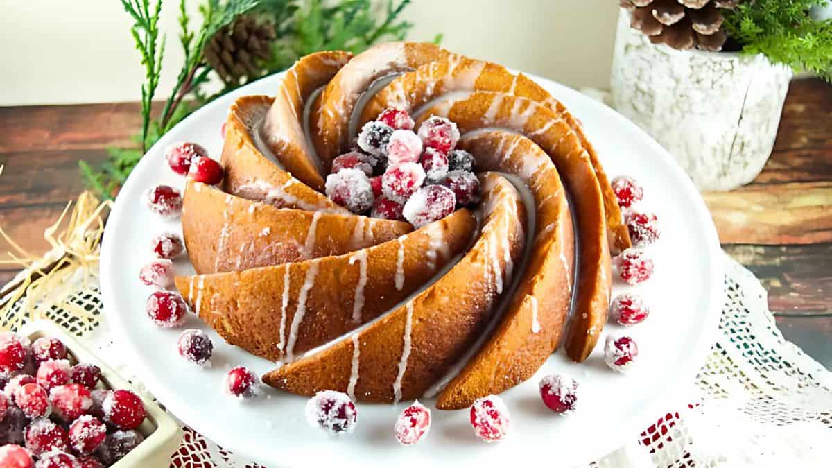 A spiral-shaped Bundt cake with icing drizzles, garnished with sugared cranberries, displayed on a white plate. Pine branches and pinecones decorate the background, enhancing the festive presentation.