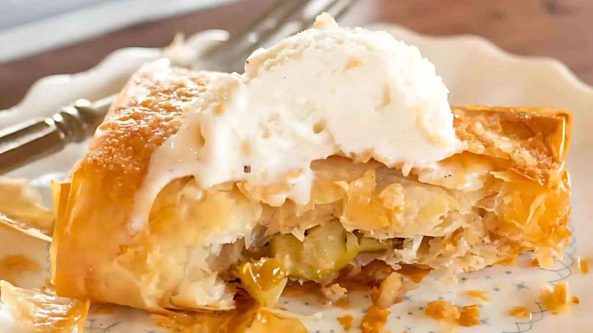 A slice of apple strudel with a flaky golden crust is topped with a scoop of melting vanilla ice cream, served on a decorative plate with a fork. The pastry filling is visible, showcasing chunks of apple.