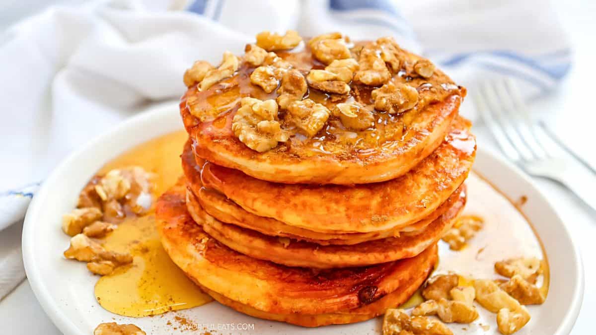 A stack of pancakes topped with chopped walnuts and drizzled with syrup sits on a white plate. A fork and a cloth napkin are visible in the background.