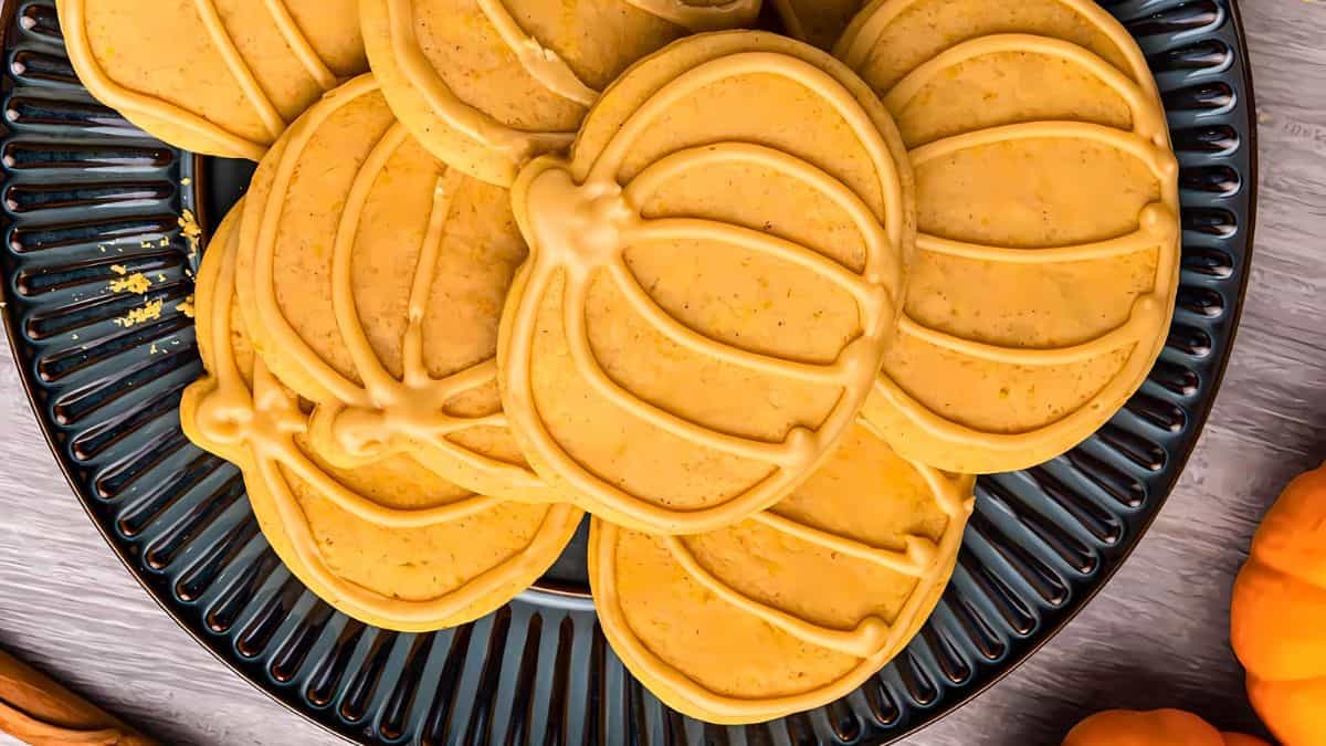A plate of pumpkin-shaped cookies with orange icing detailed like pumpkins. The cookies are arranged on a dark, decorative plate. The setting includes small pumpkins in the background.