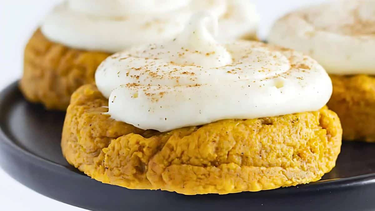 A close-up of pumpkin cookies topped with swirls of white frosting and a sprinkle of cinnamon, displayed on a black plate.