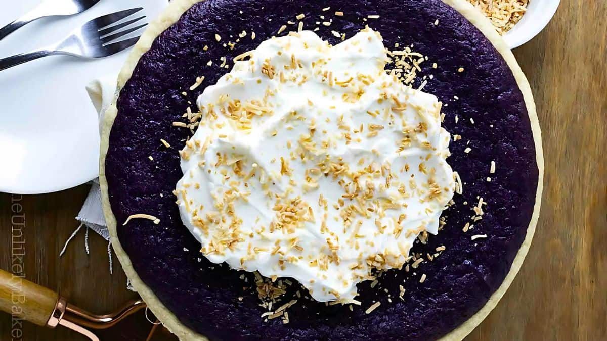 A vibrant purple yam pie topped with whipped cream and sprinkled with toasted coconut flakes. A fork, knife, and wooden serving utensils are placed beside the pie on a wooden table.