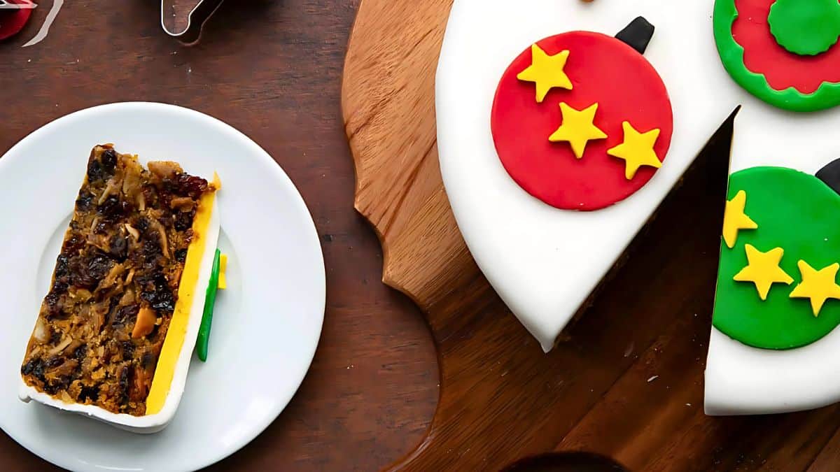 A slice of fruitcake on a white plate sits beside a round fruitcake, which is decorated with white icing and colorful Christmas ornament designs with stars. The cake is on a wooden surface.