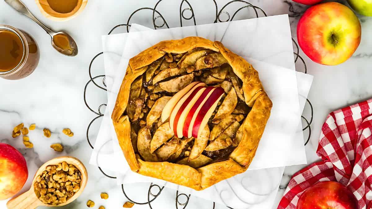 A rustic apple galette topped with thin apple slices on parchment paper. It's surrounded by fresh apples, a jar of caramel, walnuts, a spoon, and a red-checkered cloth on a marble surface.