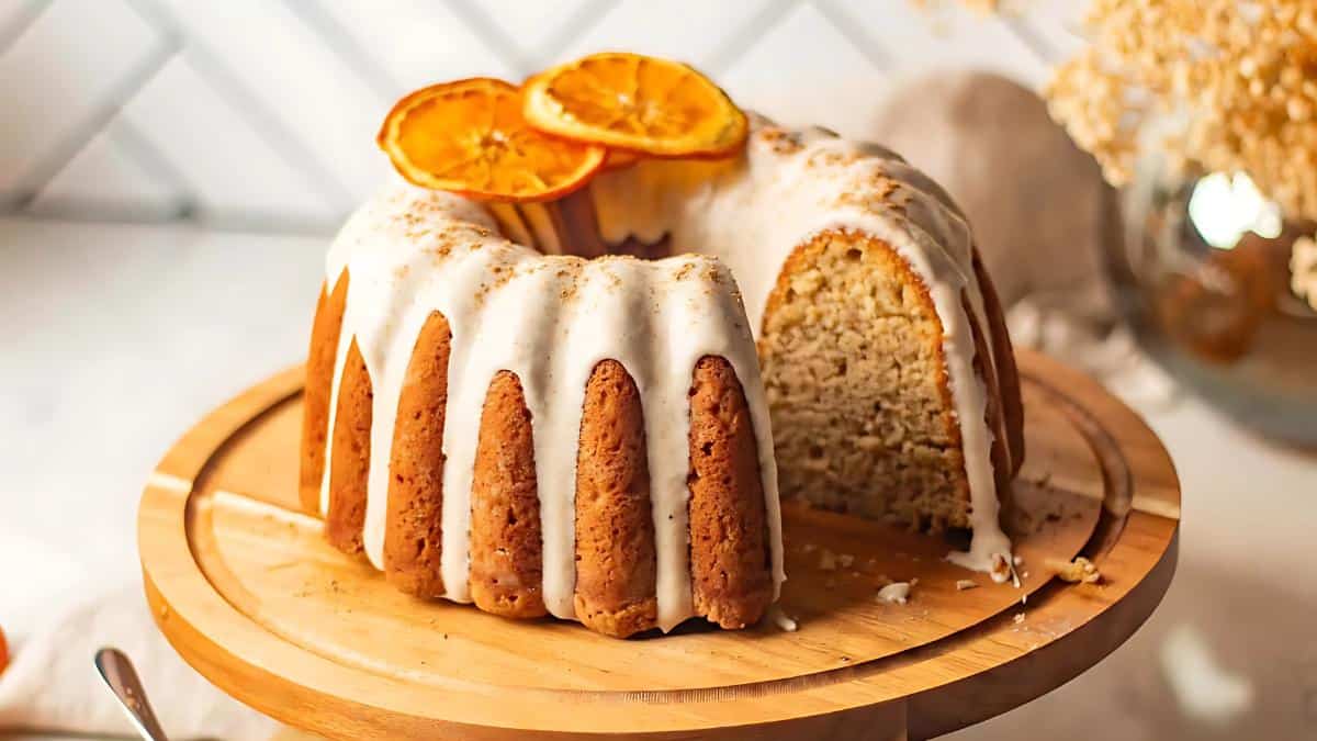 A Bundt cake with white glaze on a wooden cake stand, garnished with orange slices. A slice is missing, revealing the moist texture inside. Dried flowers and a fabric napkin are in the blurred background.
