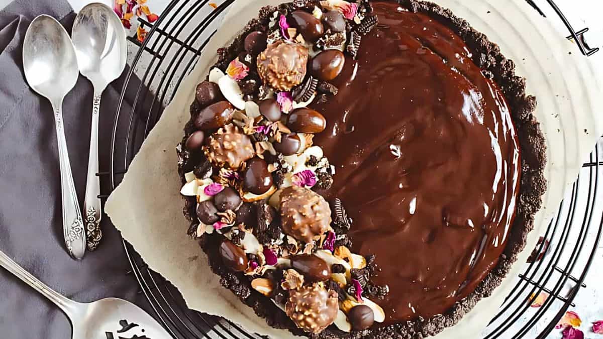 A chocolate tart with a glossy finish, garnished on one side with chocolate truffles, nuts, and colorful edible flowers, is displayed on a wire rack. Accompanying it are two ornate silver teaspoons and a dark napkin.