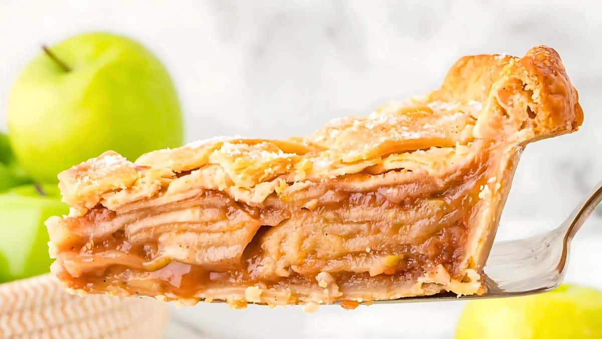 A close-up of a slice of apple pie on a spatula, showcasing layers of apples and a flaky golden crust. Green apples are blurred in the background, highlighting the fresh ingredients.