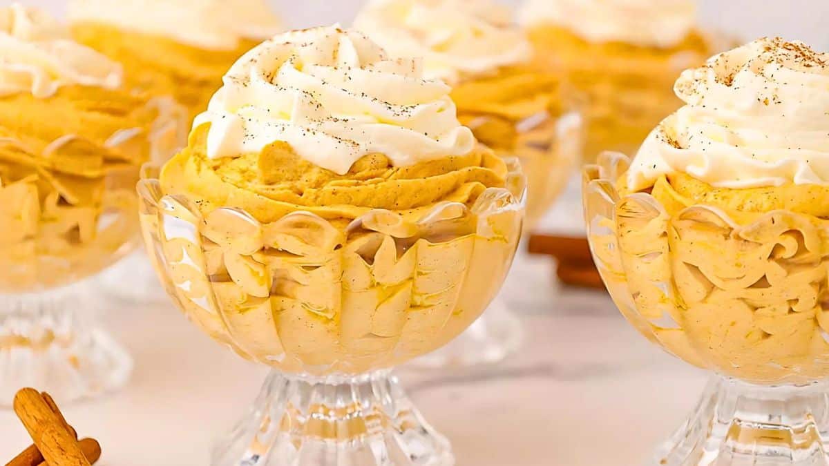 Glass bowls filled with creamy pumpkin mousse, topped with whipped cream and a sprinkle of spices, displayed on a table. Cinnamon sticks are placed nearby, adding a decorative touch to the autumnal dessert setting.