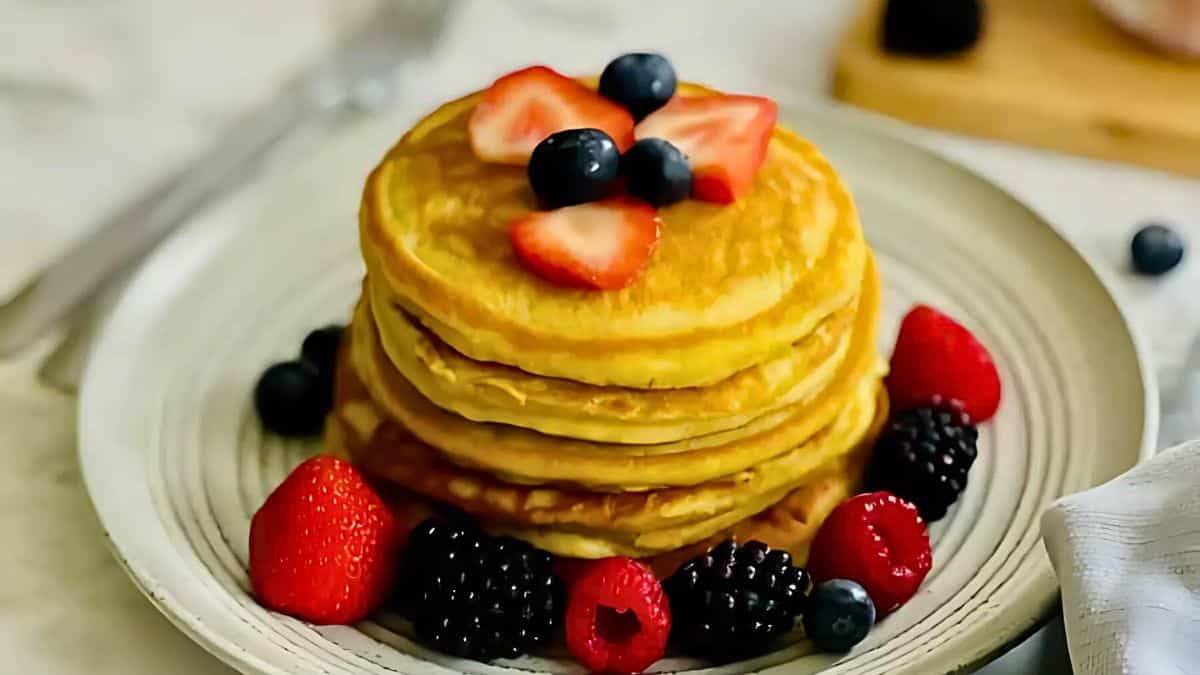 A stack of golden pancakes topped with sliced strawberries and blueberries sits on a white plate. The pancakes are surrounded by fresh raspberries and blackberries. A fork and napkin are placed nearby.