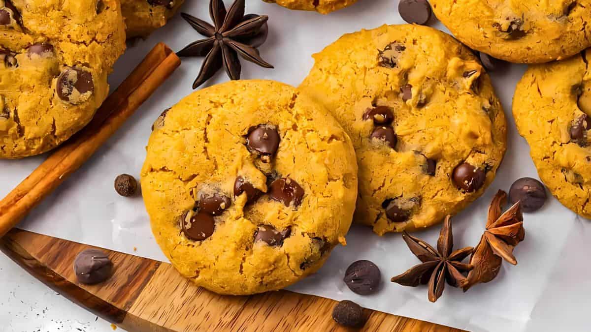 A close-up of pumpkin chocolate chip cookies on a wooden board. The cookies are surrounded by whole star anise, cinnamon sticks, and chocolate chips scattered around, creating a cozy, autumnal setting.