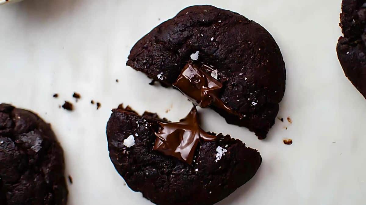 A chocolate cookie with a gooey, melted chocolate center is broken in half. The cookie appears dark and moist, with a few grains of sea salt sprinkled on top, set against a plain white background.