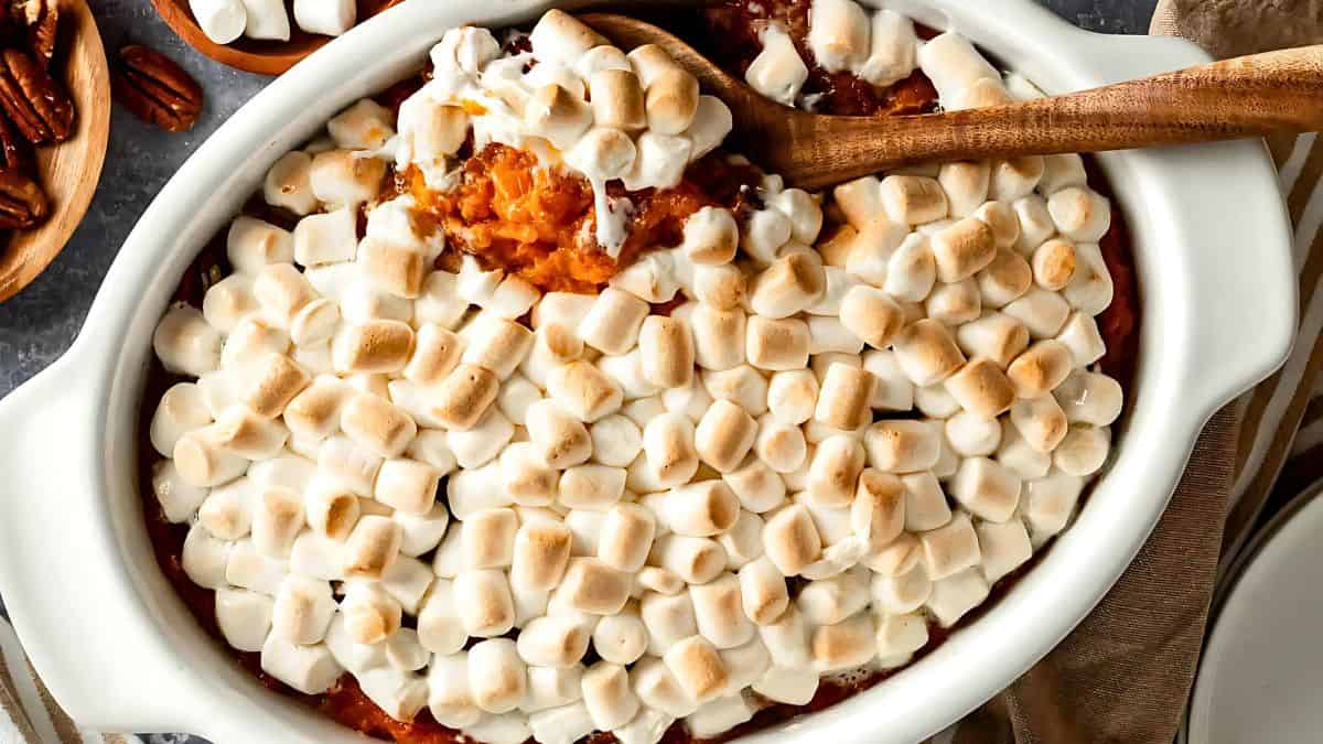 A casserole dish filled with sweet potato casserole topped with toasted marshmallows. A wooden spoon is scooping some of the casserole, revealing the orange sweet potato underneath.