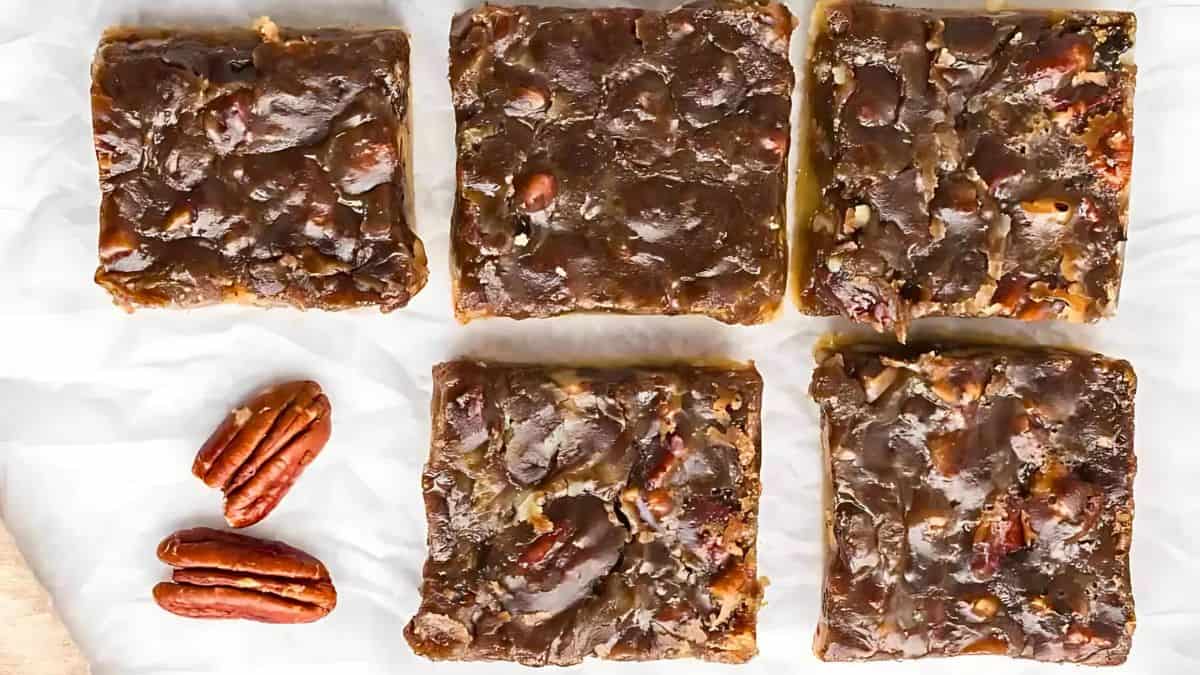 Five square pecan pie bars are arranged on a white surface with three pecans placed beside them. The bars have a glossy, nutty topping.