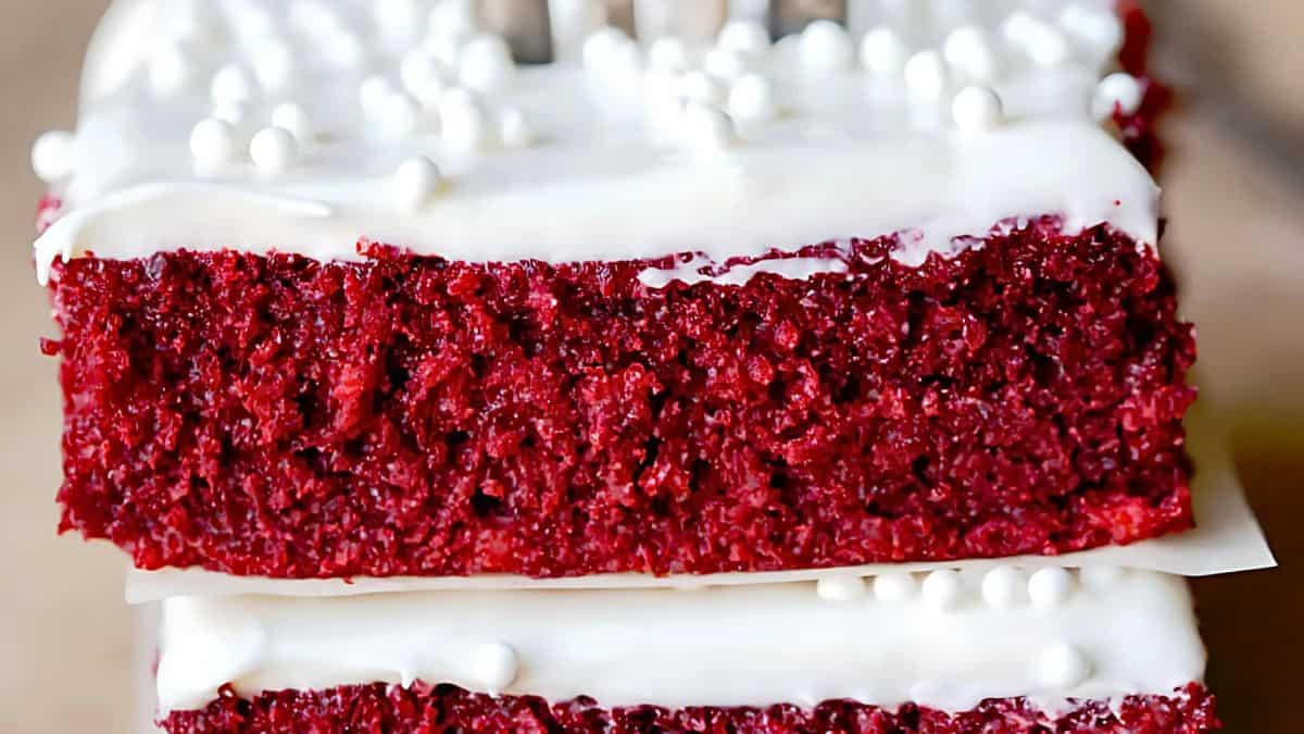 Close-up of two slices of red velvet cake stacked on top of each other, topped with white frosting and small white sprinkles. A fork is partially visible behind the slices.