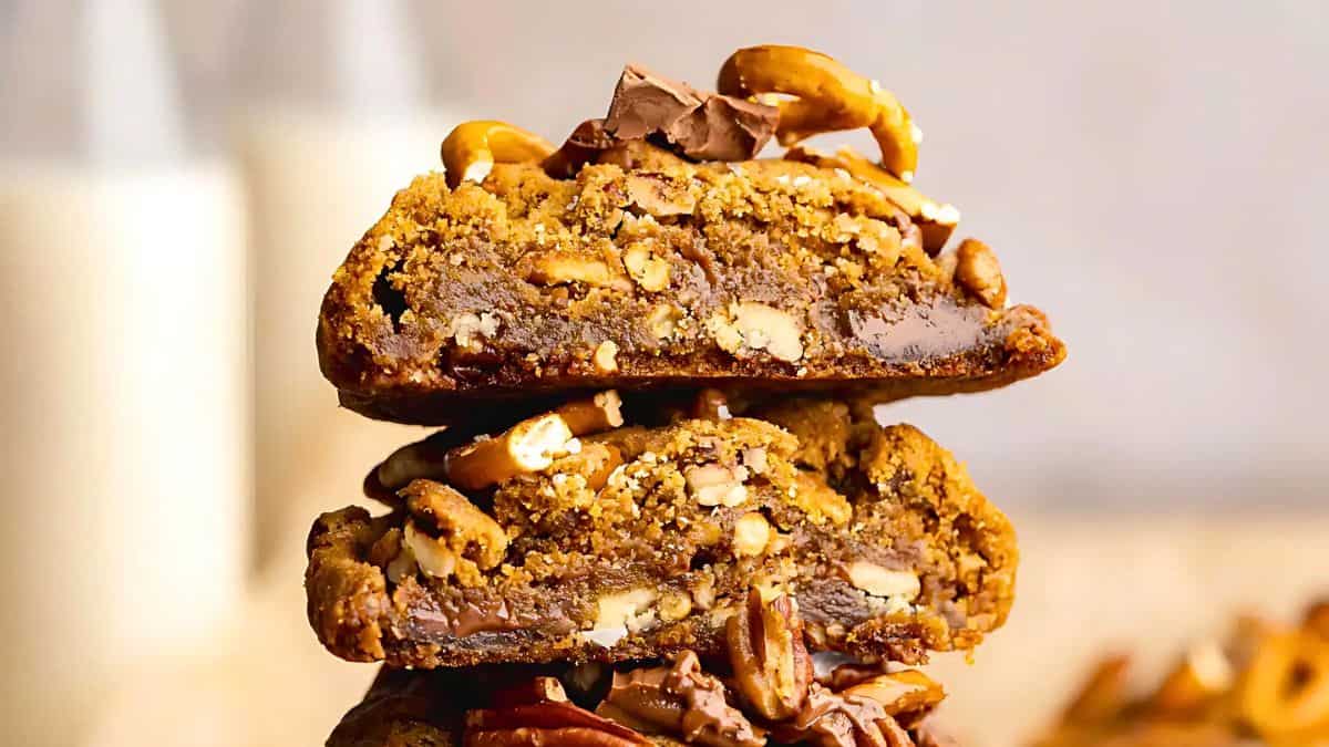 A close-up of two chocolate chip cookies stacked on top of each other. The cookies are thick, filled with chunks of chocolate and nuts, and have a soft, crumbly texture. Some pretzel pieces are visible on the top cookie.