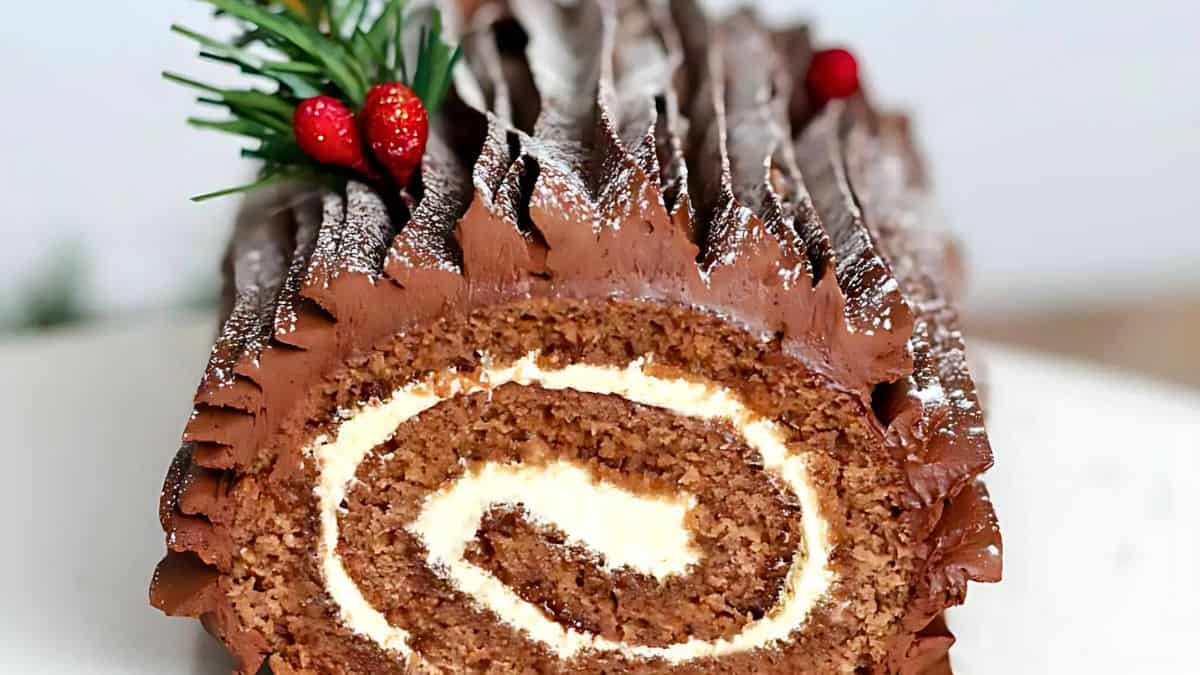 A delicious yule log cake with chocolate frosting and a creamy swirl is decorated with small red berries and green leaves. The cake has a textured surface with powdered sugar dusting.