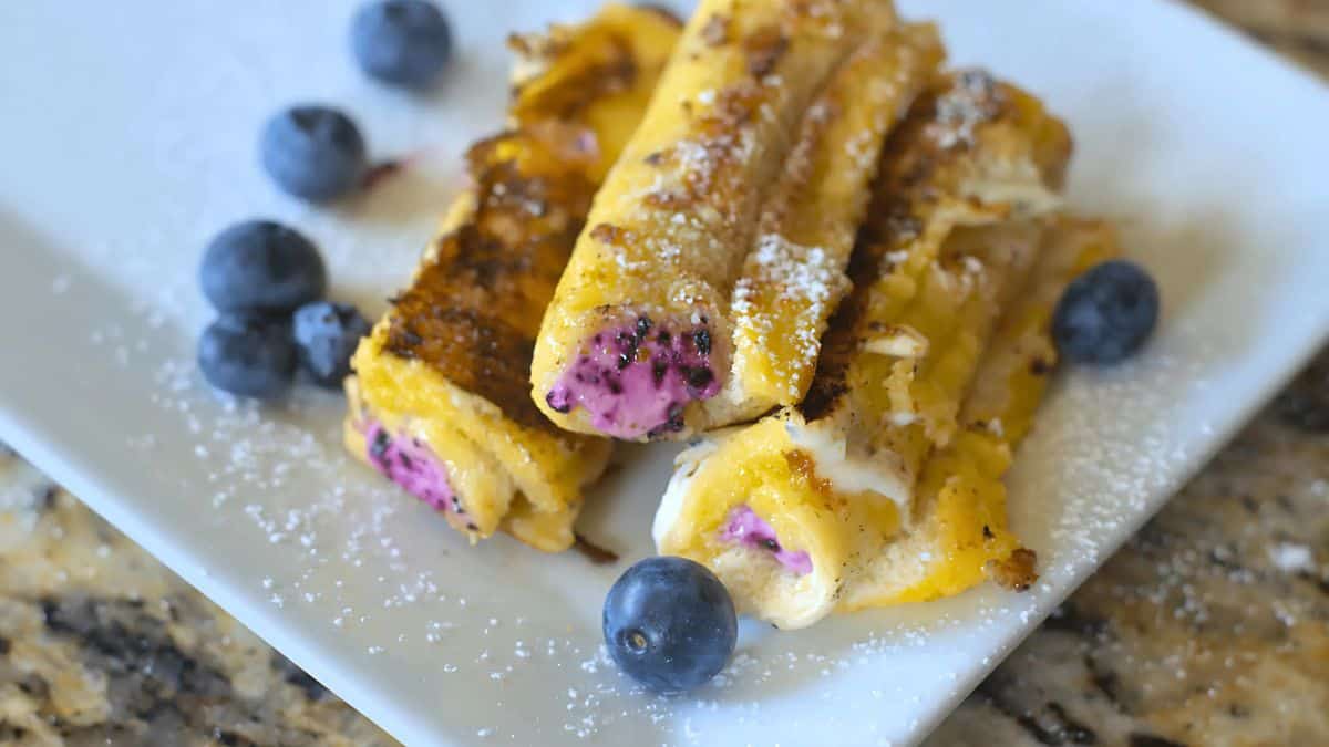 Close-up of three rolled crepes filled with a pink berry cream and garnished with fresh blueberries. The crepes are lightly dusted with powdered sugar and served on a white rectangular plate on a marble countertop.