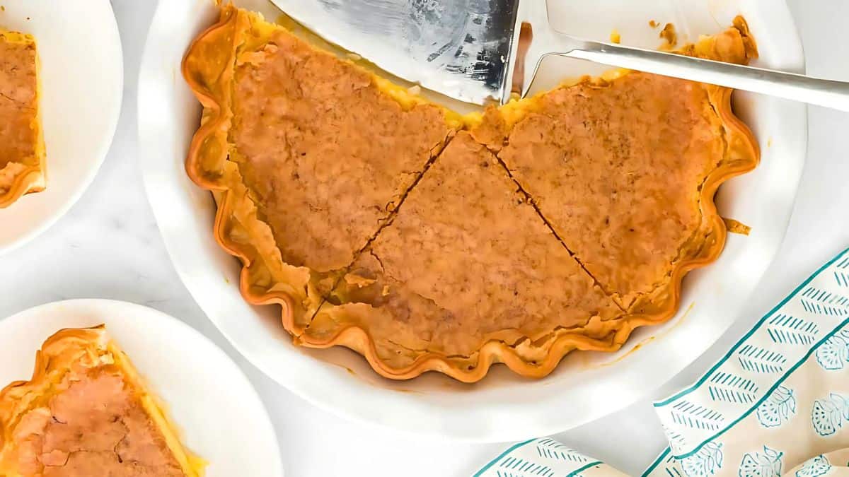 A close-up of a pie with a golden brown crust, partially sliced into pieces, on a white plate. A metal pie server rests nearby. Additional slices are on smaller plates, and a patterned cloth is visible in the corner.