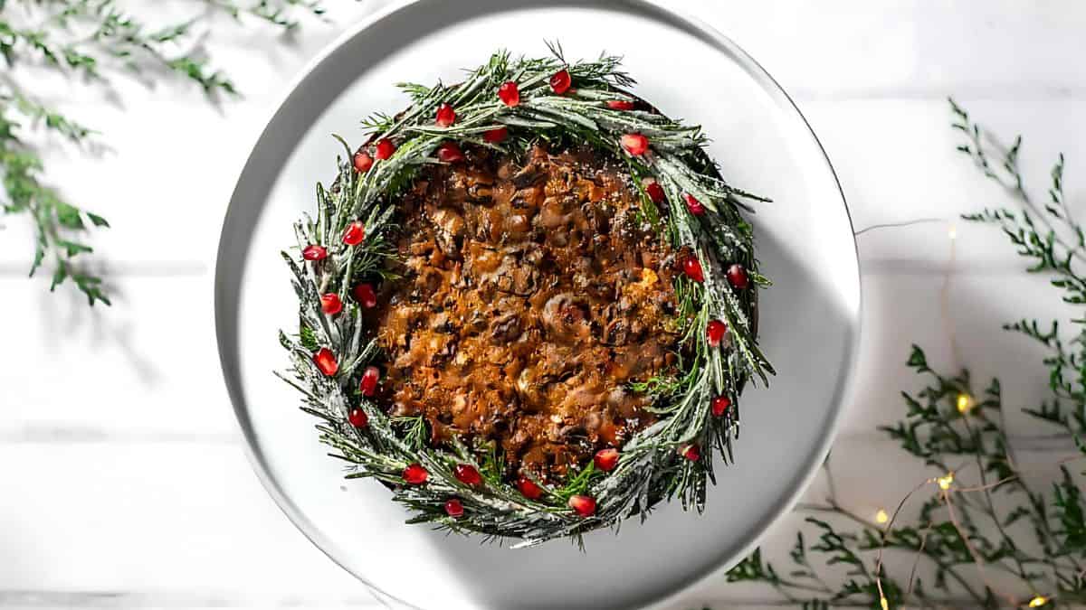 A round fruitcake on a white plate, adorned with a festive wreath of rosemary sprigs coated in sugar and decorated with red cranberries. The background features scattered greenery on a white surface.