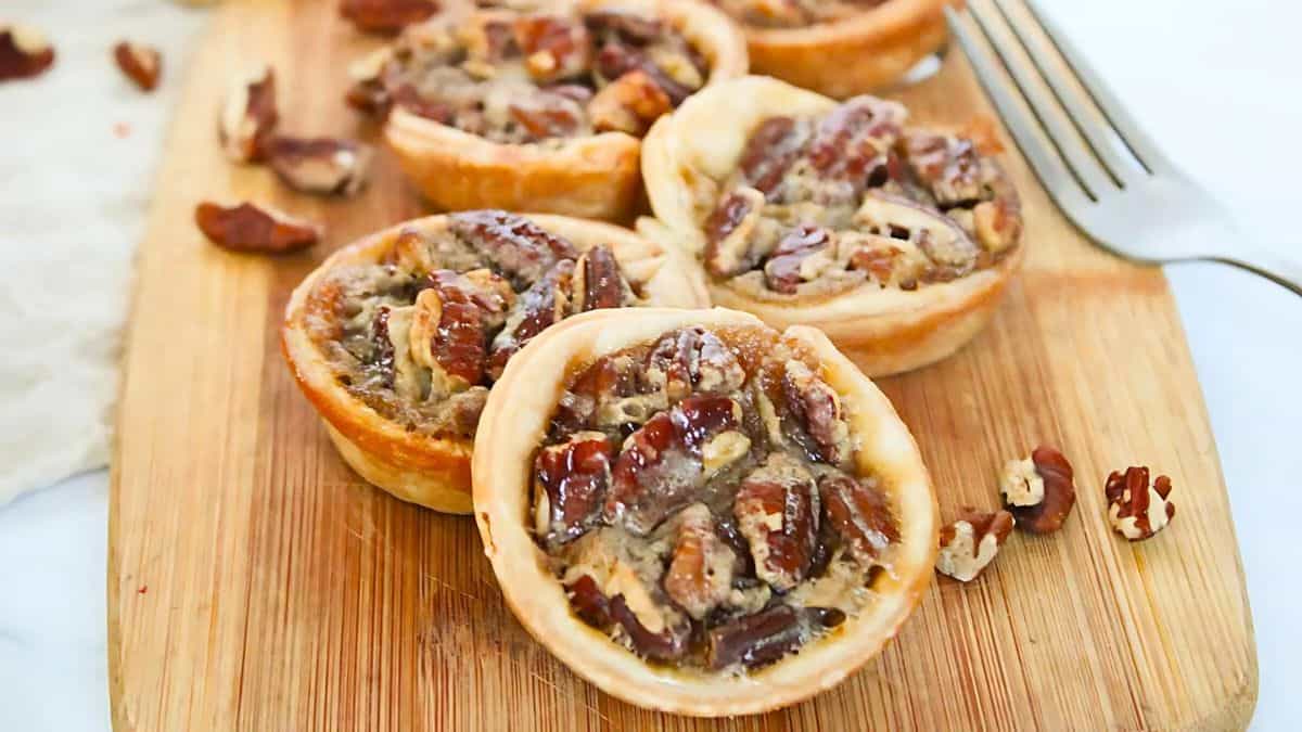 Close-up of pecan tarts on a wooden board. The tarts have a flaky crust and are filled with a gooey pecan mixture. Scattered pecan pieces and a fork are nearby.