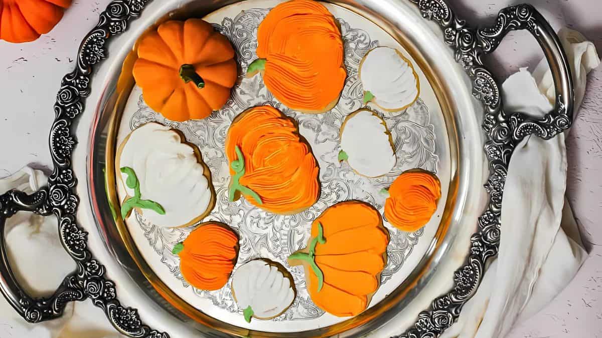 A decorative silver tray holds cookies shaped like pumpkins in orange and white, adorned with green accents. The cookies rest on an ornate surface, and the entire setup is placed on a soft, white fabric backdrop.