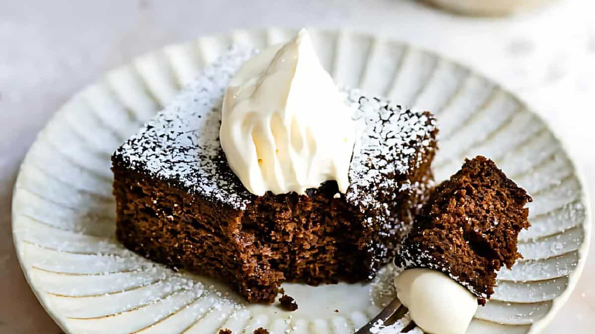 A piece of chocolate cake dusted with powdered sugar sits on a white plate. A dollop of whipped cream tops the cake, and a fork has taken a bite, resting on the plate next to the slice.
