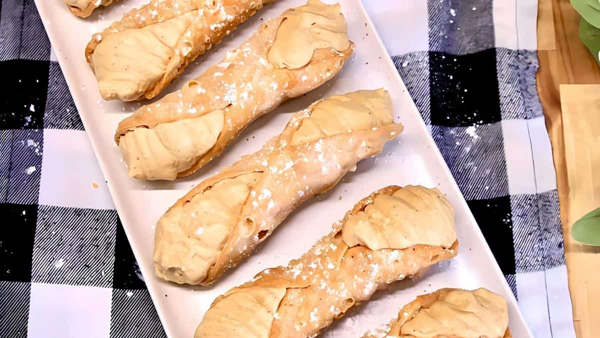 A rectangular white platter holds five crispy cannoli, filled with a creamy beige filling and dusted with powdered sugar, resting on a black and white checkered tablecloth.
