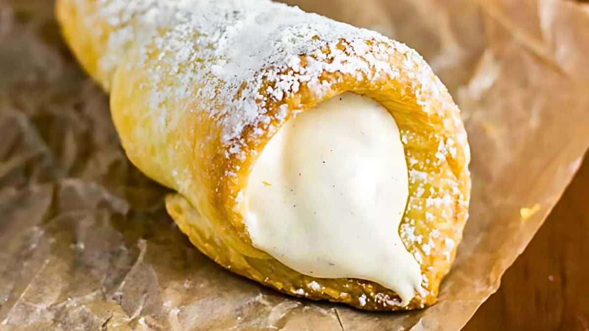 A close-up of a cream horn pastry on brown parchment paper. The flaky pastry is dusted with powdered sugar and filled with a creamy white filling.