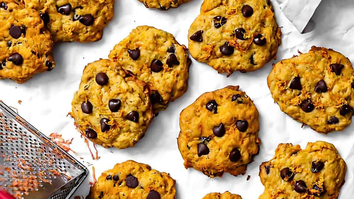 A spread of freshly baked chocolate chip cookies on a white parchment surface, with a cheese grater on the left. The cookies are golden brown, with visible chocolate chips.