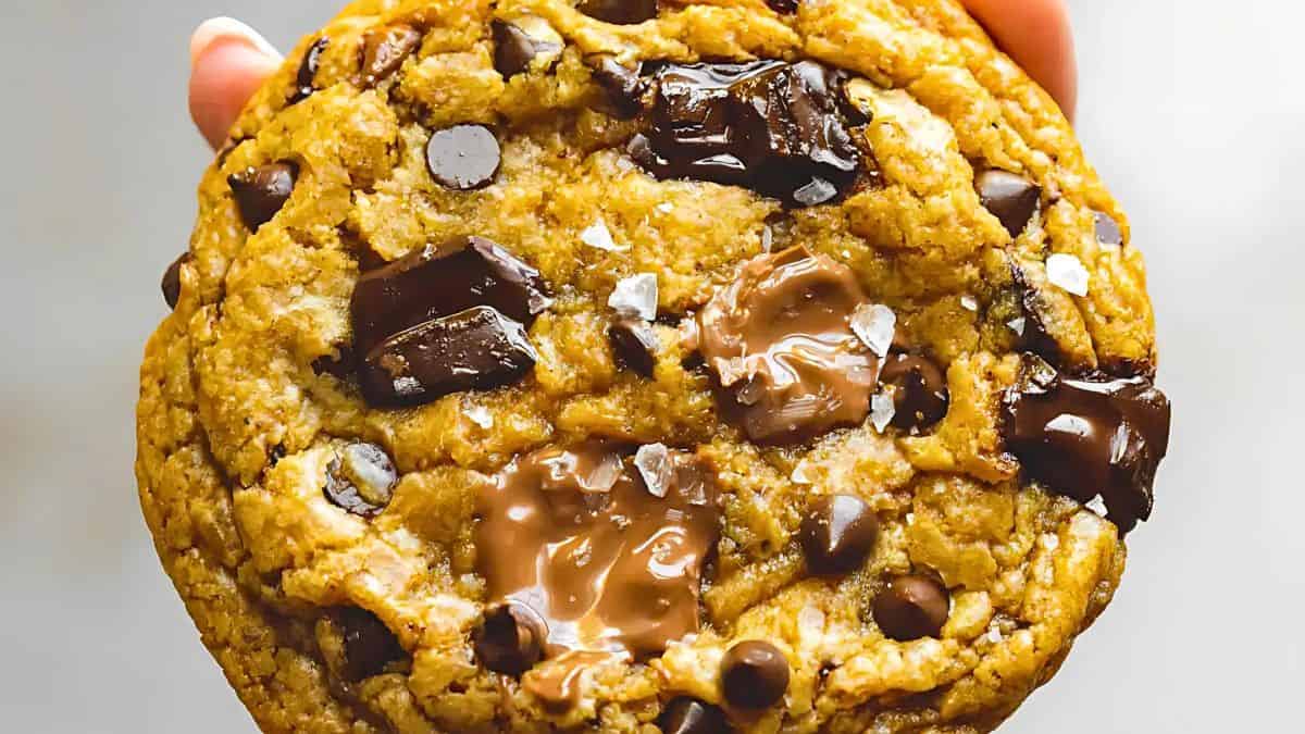 A hand holding a large chocolate chip cookie with chunks of melted chocolate dispersed throughout. The cookie has a golden-brown color and a slightly soft, gooey texture, with visible chocolate pieces and sea salt flakes on the surface.