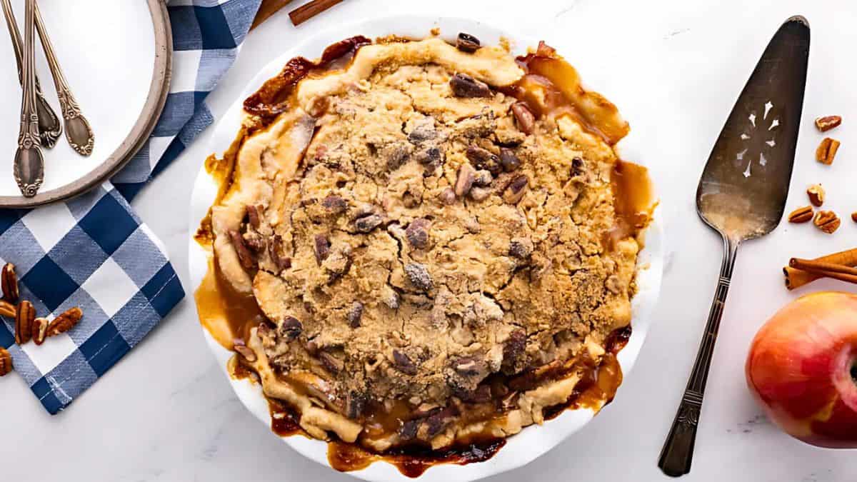 A freshly baked apple crisp topped with a golden-brown crumb topping and pecans, surrounded by a dessert server, apple, cinnamon sticks, and a plate with spoons on a checkered cloth. The dish has caramelized edges and a rustic appearance.