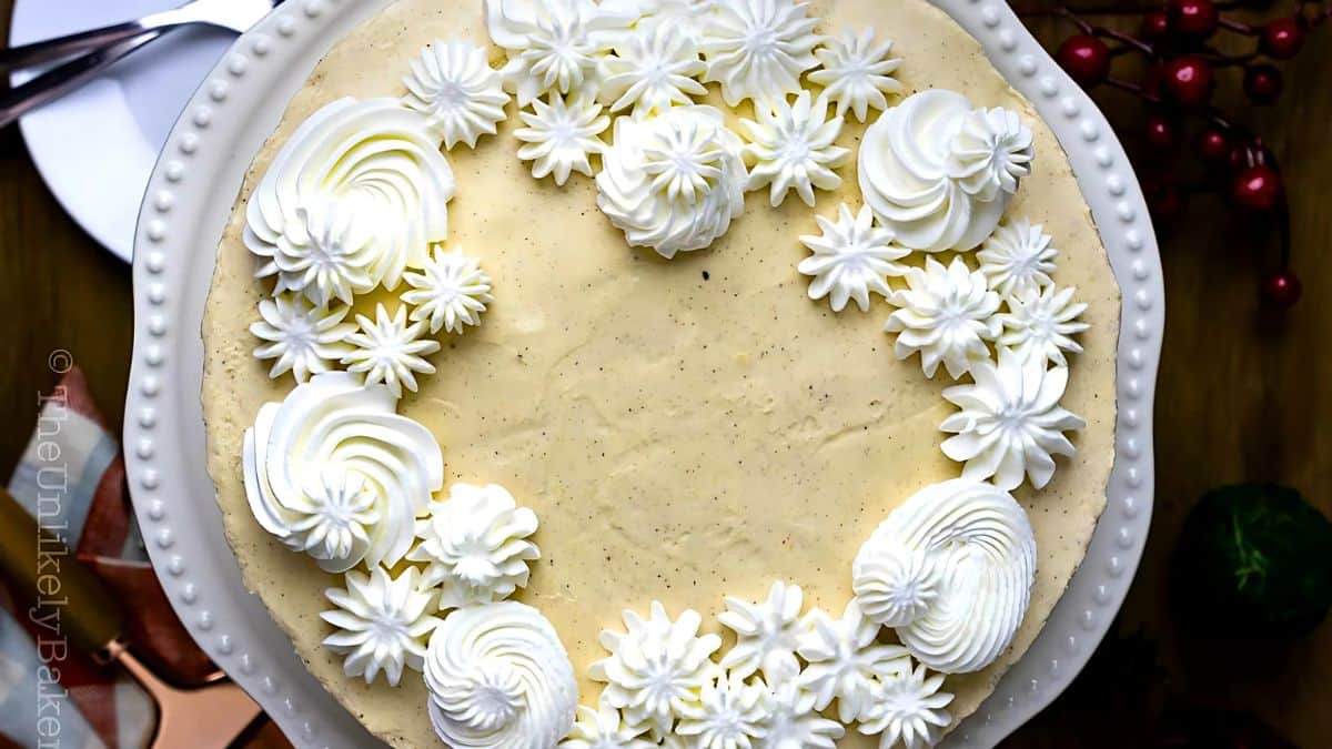 A cake with creamy frosting, topped with decorative white swirls and star-shaped patterns, placed on a white plate. Nearby, there are cherries and a dark green object partially visible.