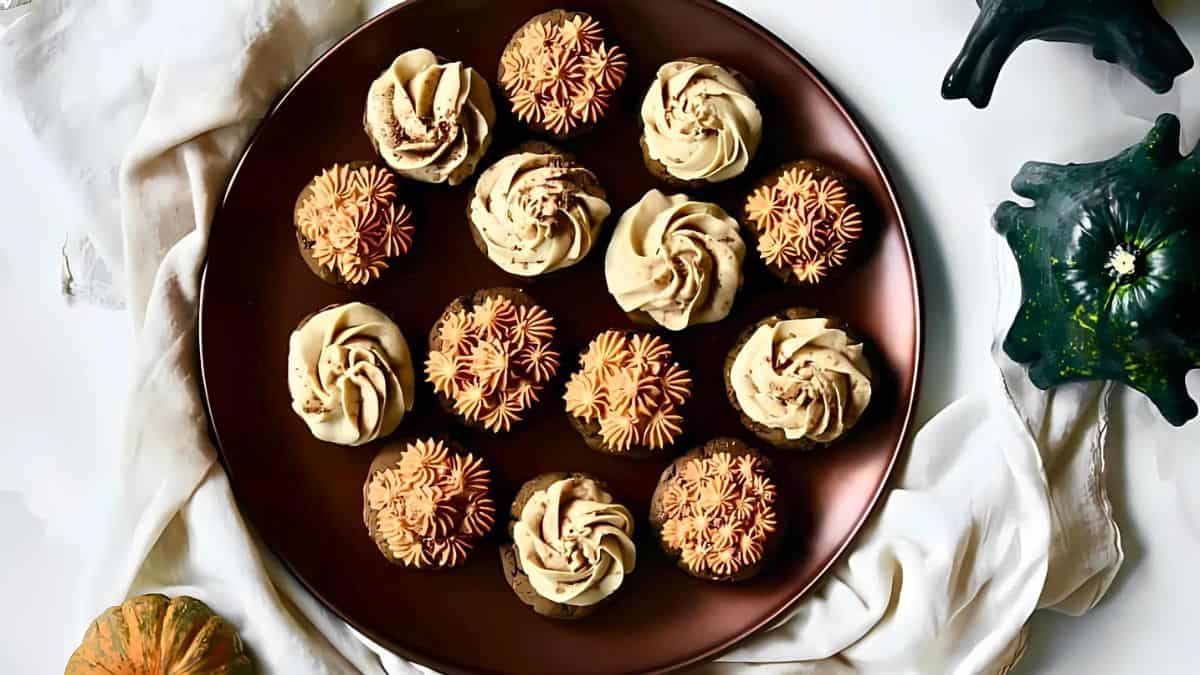 A round brown plate with cupcakes topped with beige and pinkish-orange frosting, arranged in a pattern. The plate is on a white cloth, surrounded by small decorative pumpkins and gourds.
