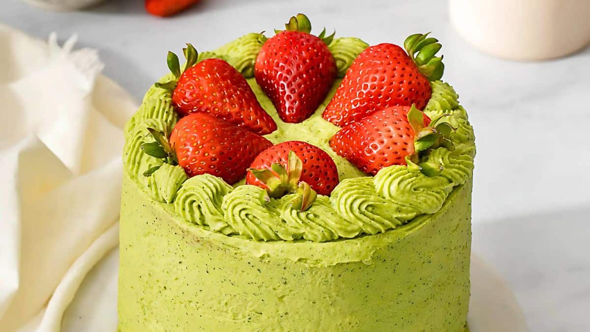 A round cake with vibrant green frosting, topped with fresh strawberries. The frosting is piped decoratively around the edge, and the cake is set on a white surface with a white cloth partially visible in the background.