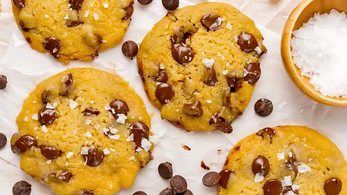 Five freshly baked chocolate chip cookies sprinkled with flaky sea salt are arranged on a parchment paper. Chocolate chips are scattered around, and a small wooden bowl filled with salt is visible in the top right corner.