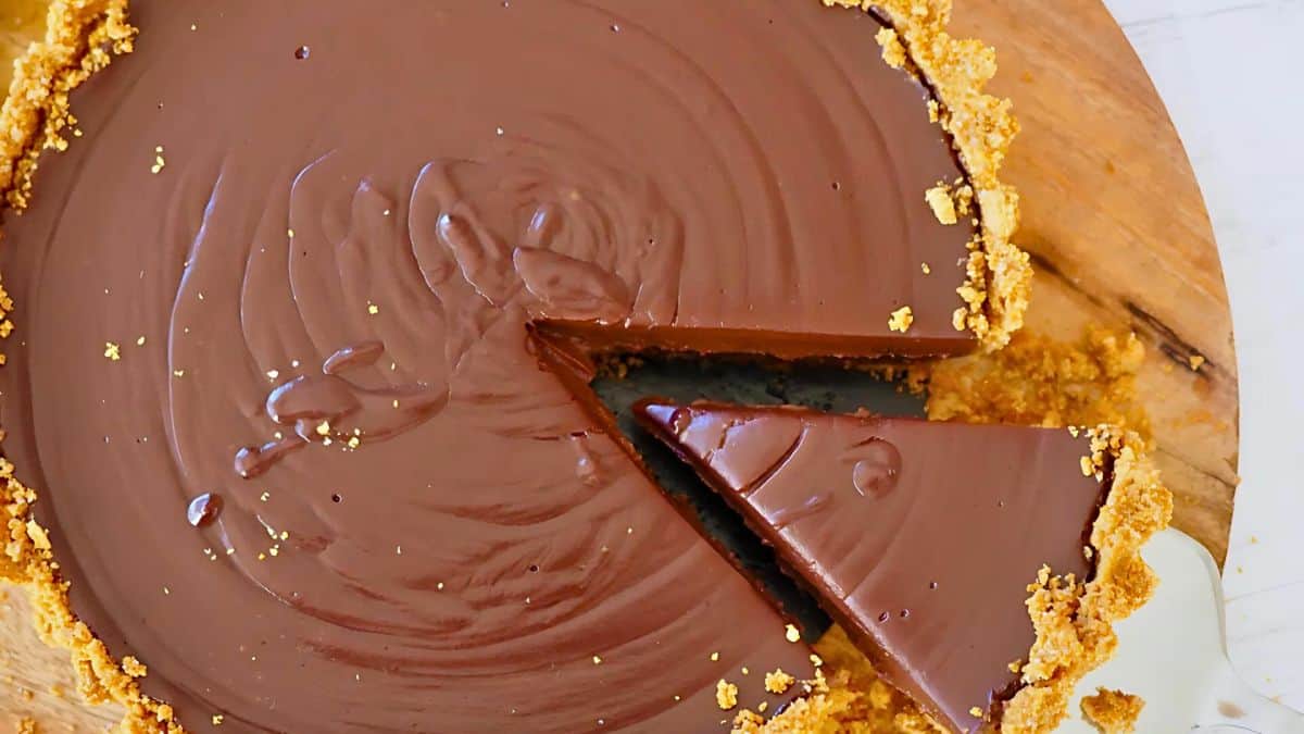 A top view of a rich chocolate tart with a smooth surface and a golden crust. One slice is partially removed, revealing the glossy interior, on a wooden cutting board.