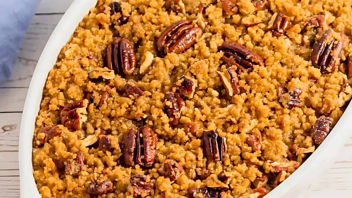 A close-up of a baked sweet potato casserole in a white dish. It is topped with a crumbly brown sugar and pecan mixture, giving it a textured, golden-brown appearance. The dish is placed on a light-colored wooden surface.