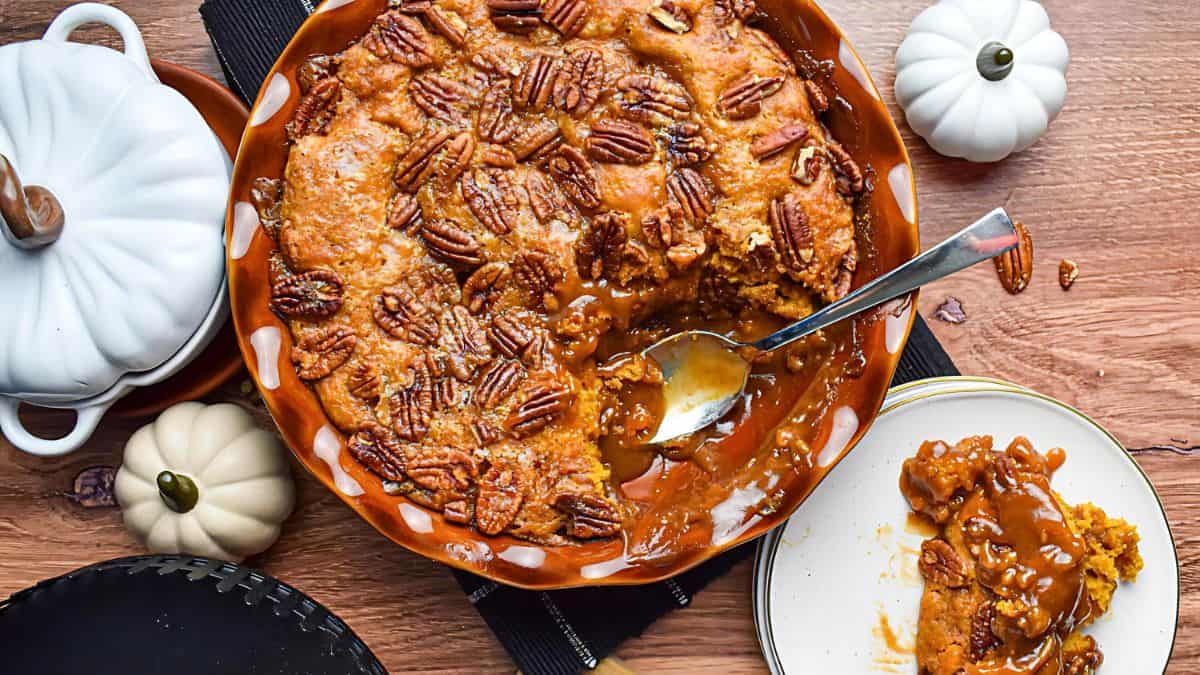 A pecan-topped dessert in a brown pie dish with a portion served on a white plate. A spoon rests in the dish. Two decorative white ceramic pumpkins and small real pumpkins are nearby on a wooden surface.