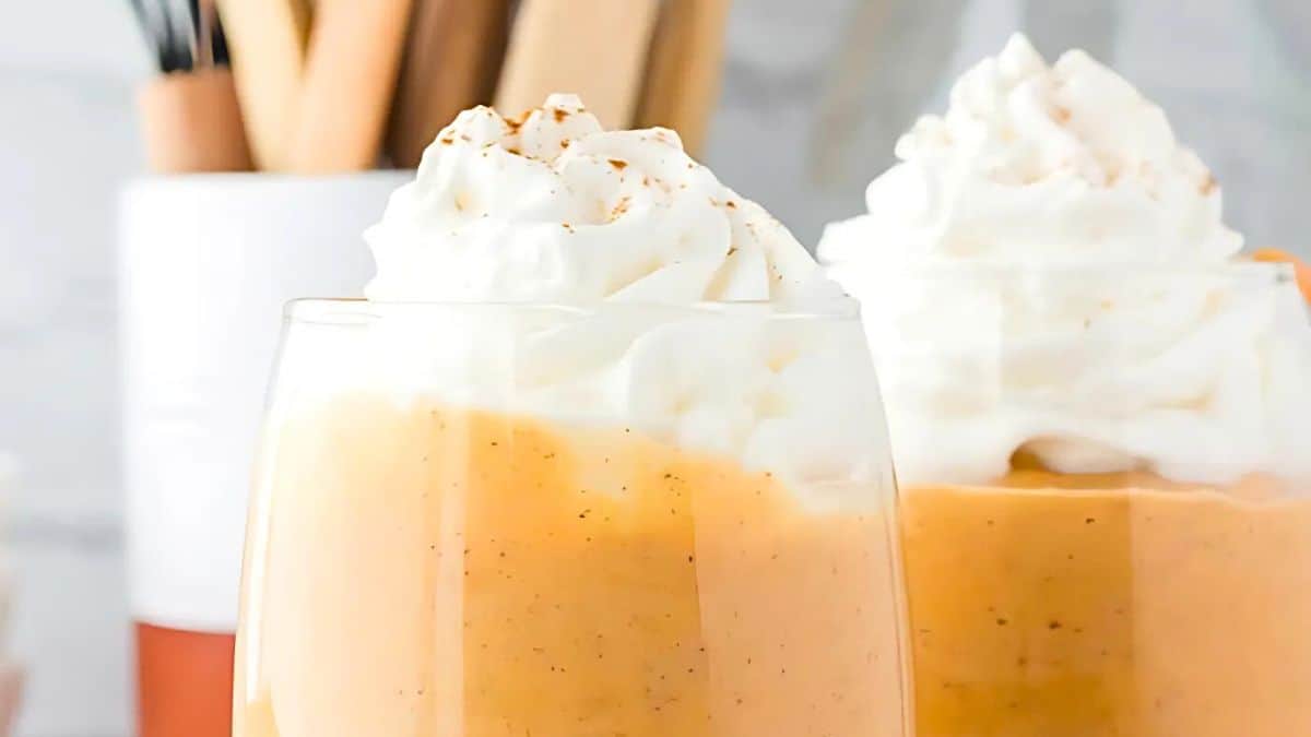 Two clear glasses filled with creamy pumpkin mousse topped with swirls of whipped cream and a sprinkle of spice. The background features kitchen utensils in a container.