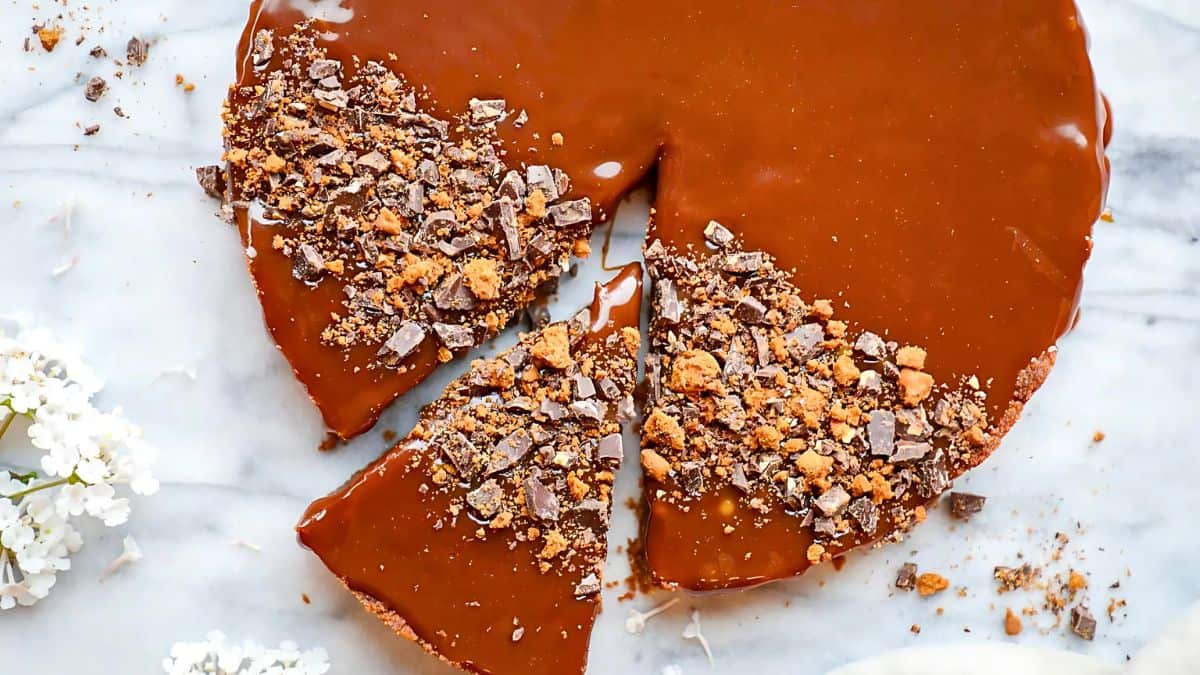 A round caramel pie on a marble surface, topped with crushed chocolate pieces. One slice is cut and slightly separated from the rest, showcasing the rich caramel topping. White flowers adorn the corner of the image.