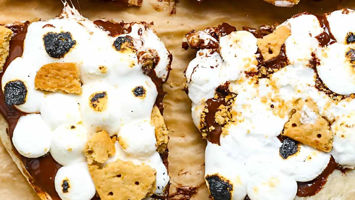Close-up of toasted marshmallows and pieces of graham cracker on melted chocolate, mimicking a s'mores dessert. The ingredients are set on a parchment paper background, with marshmallows showing a golden-brown, slightly charred texture.
