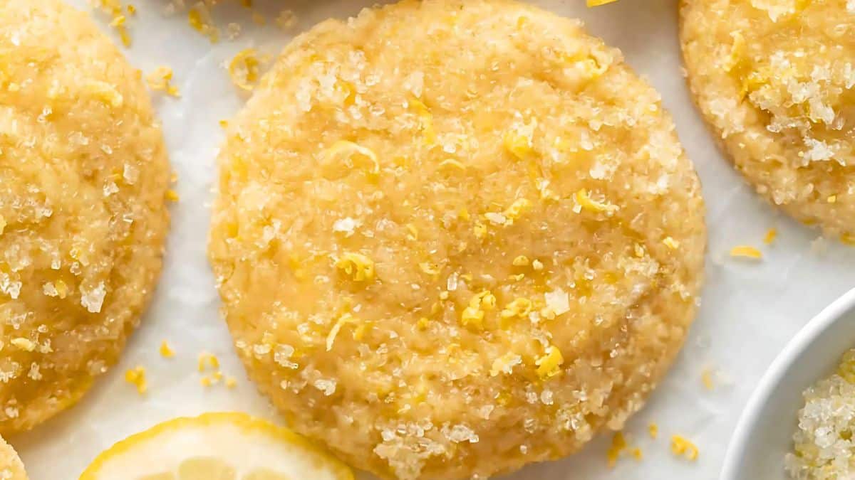 Close-up of a lemon cookie topped with coarse sugar and lemon zest, surrounded by more cookies and a lemon slice. The cookie's texture appears soft and crumbly, with a golden-yellow color.