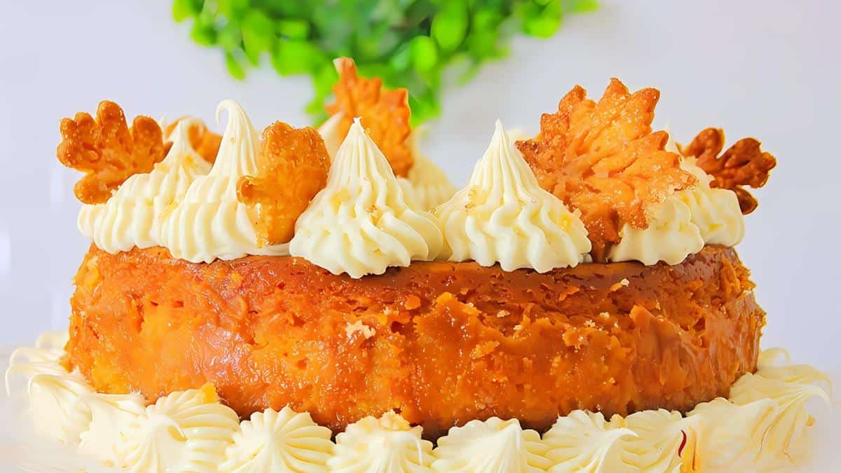 A pumpkin spice cake garnished with cream swirls and leaf-shaped cookies, set against a blurred green plant background.